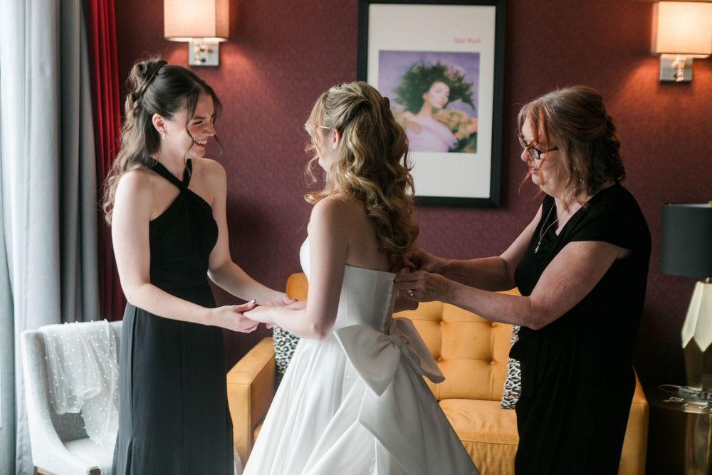 Bride being helped into white wedding gown by bridesmaid and mother in elegant hotel room.