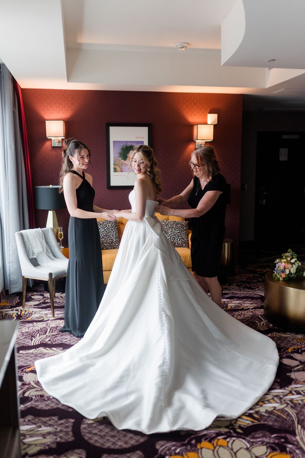 Bride in white gown being helped by two women in black dresses in an elegant hotel suite.
