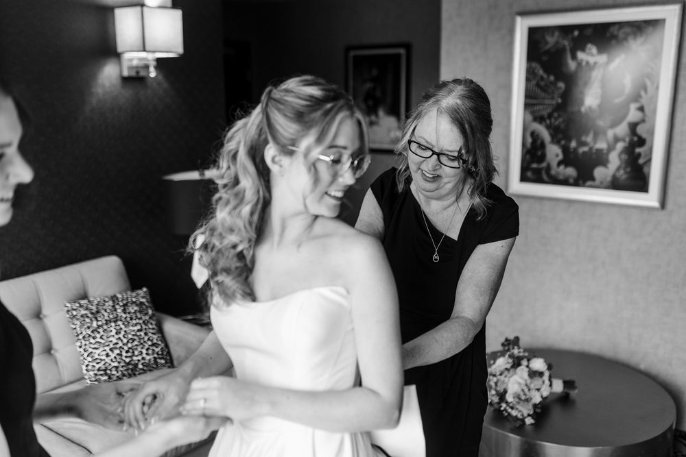 Black and white photo of a bride in a strapless gown being helped by a woman in a hotel room.