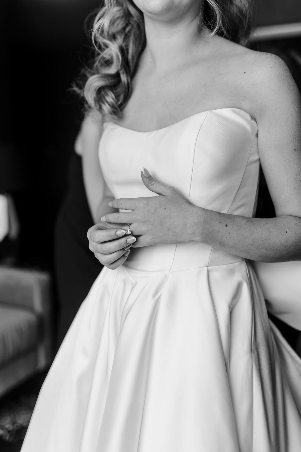 Black and white photo of a bride in a strapless satin wedding gown, with hands clasped at her waist.