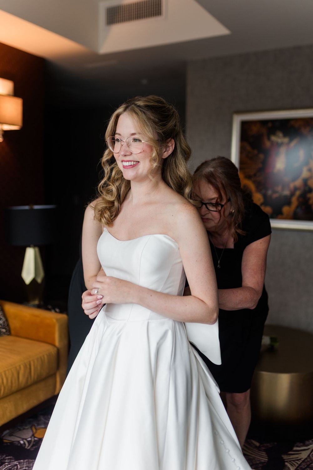 Smiling bride in strapless white gown being helped into her dress by a woman in a hotel room.