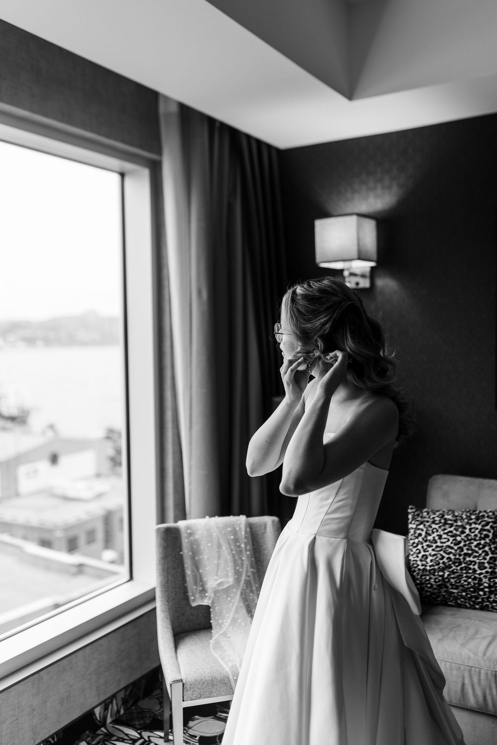 Bride in strapless white gown putting on earrings by a large window in a hotel room.