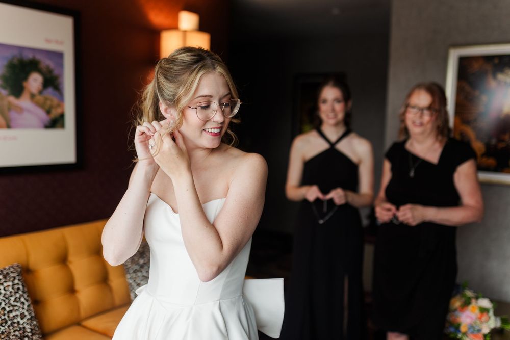 Bride in white strapless gown adjusting earrings while bridesmaids in black dresses look on in elegant hotel room.