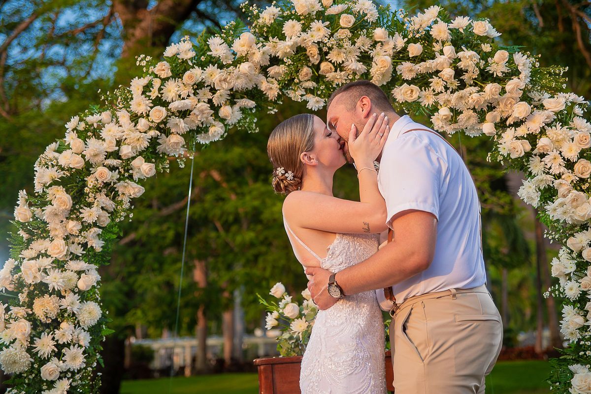 Bride in a white lace gown and groom in a white shirt and khaki pants share a kiss under a white floral wedding arch outdoors.