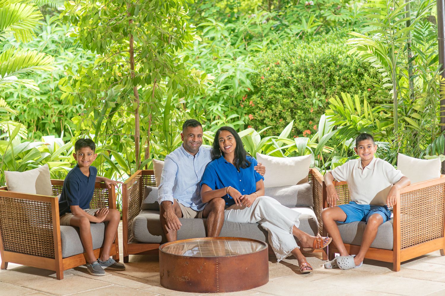 Smiling family of four relaxing on wicker lounge chairs around a round copper table on a patio surrounded by lush tropical greenery.