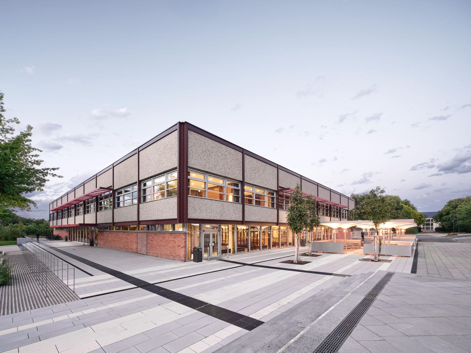 Modern two-story building with brick base and glass facade beside a paved plaza with young trees under a soft evening sky.