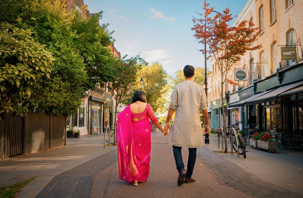 Indian couple in traditional attire holding hands walking down a sunny European street lined with trees and shops.