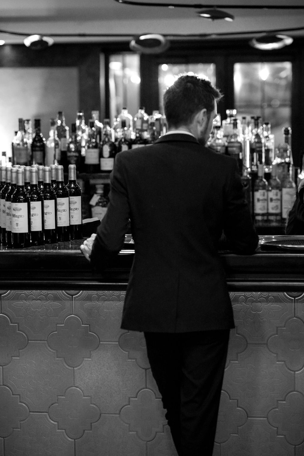 Black and white photo of a bartender in a suit standing behind a bar stocked with wine and spirit bottles.