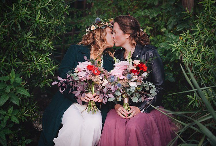Two brides kissing while holding colorful floral bouquets, surrounded by lush greenery on their wedding day.