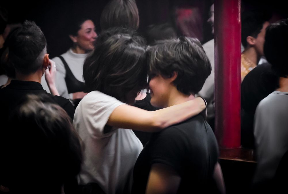 Two people embrace intimately in a crowded nightclub, surrounded by other guests in a dimly lit venue.