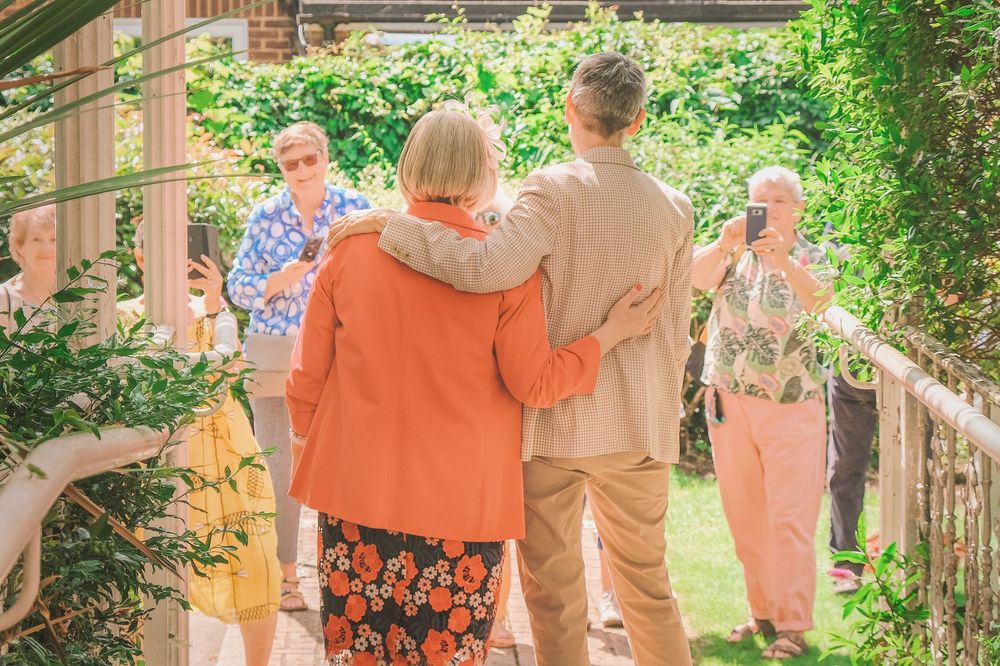 Elderly couple with arms around each other walking through a sunny garden at an outdoor social gathering.