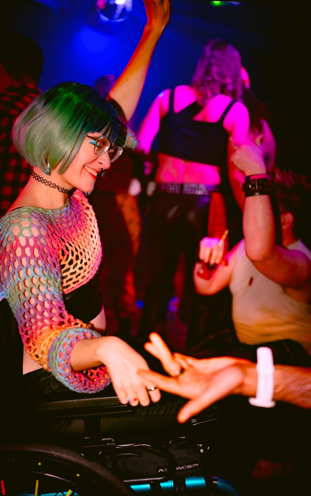A young woman with green hair in a mesh top dances joyfully in a wheelchair at a vibrant nightclub.