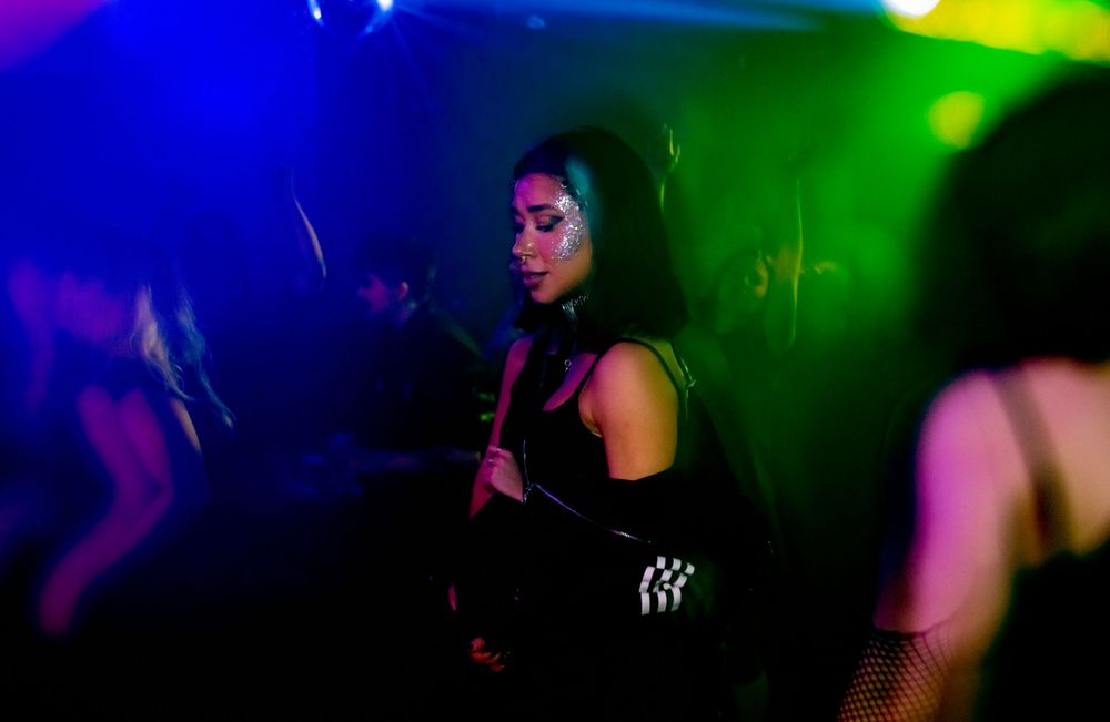A woman in a black outfit dances under colorful neon lights at a nightclub.