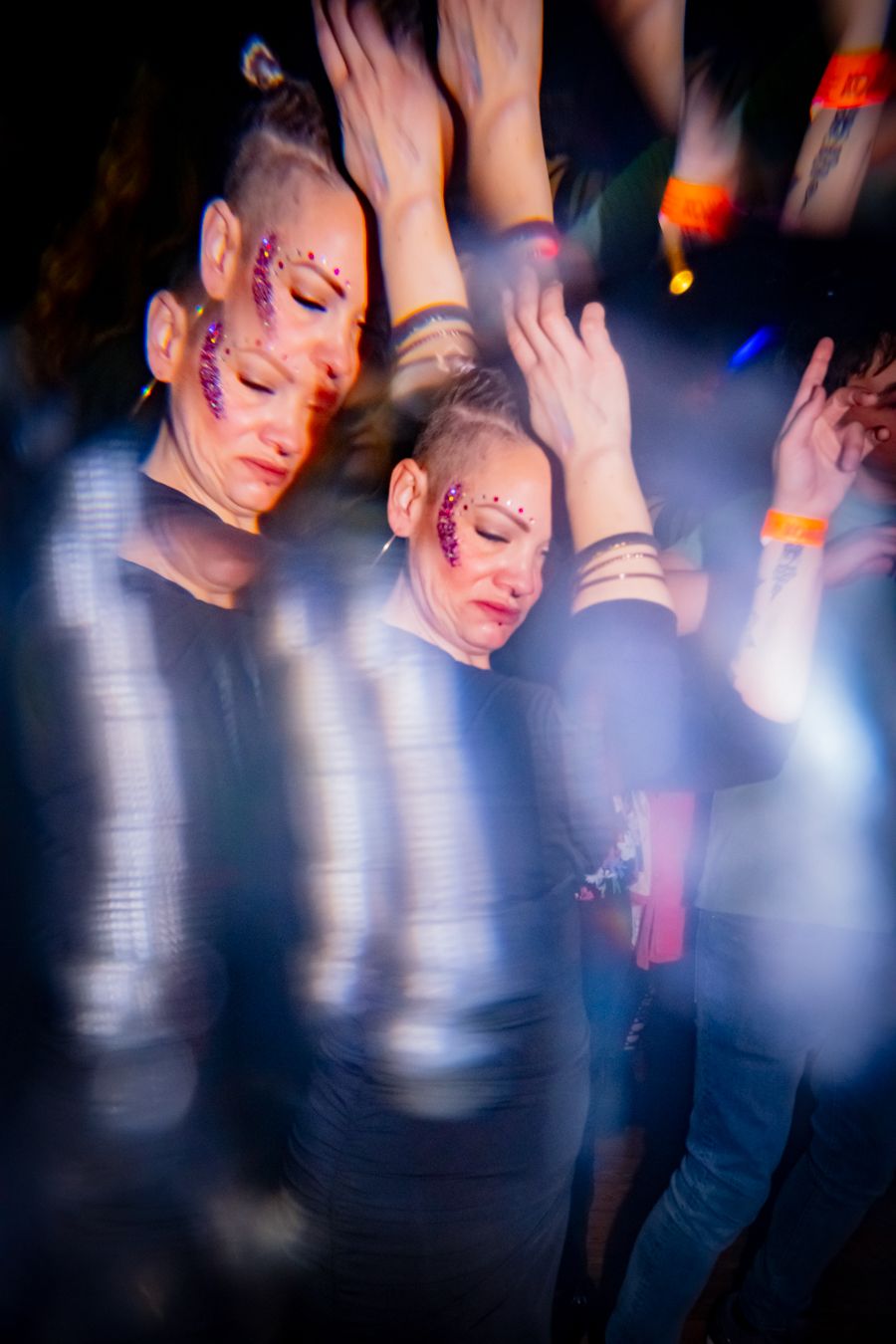 Two women with glitter face makeup dancing at a nightclub event with motion blur effects.