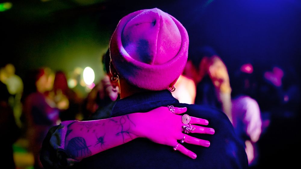 Two people embrace at a nightclub, one wearing a pink cap with tattoos visible under neon lights.