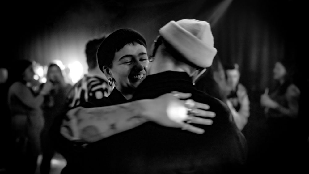 Two people share a warm embrace at a lively event, captured in black and white photography.