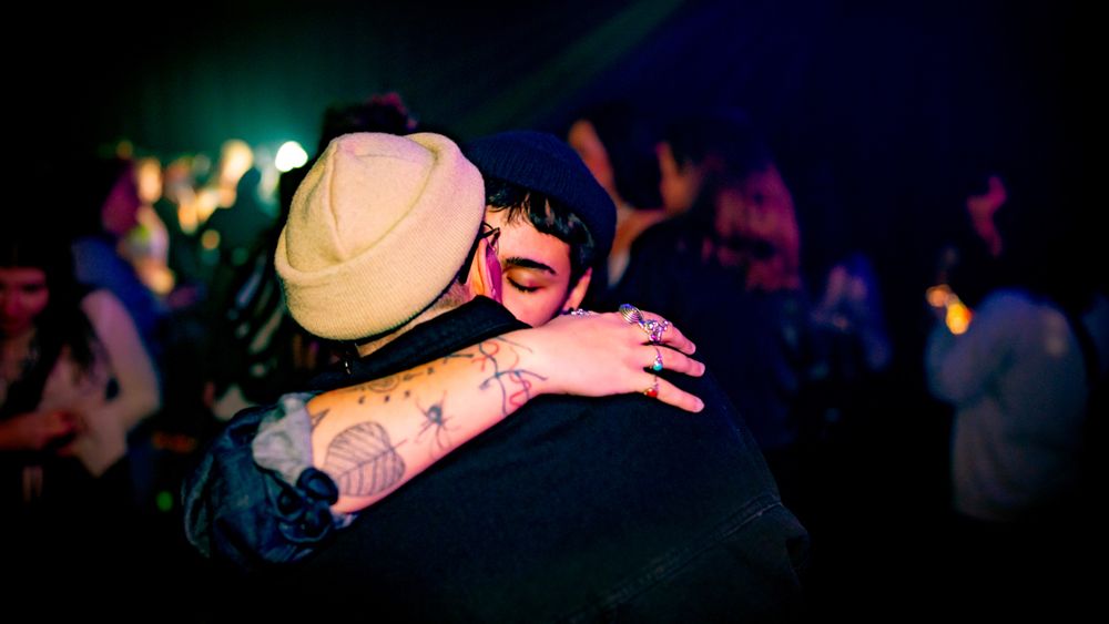 Two people share a warm hug at a nightclub, one wearing a tan hat and showing tattoos on their arm.