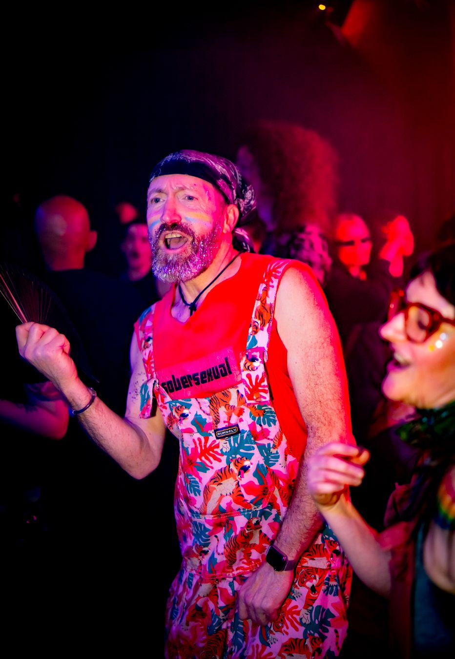 A bearded man in colorful floral overalls and red bandana dances joyfully at a vibrant nightclub event.