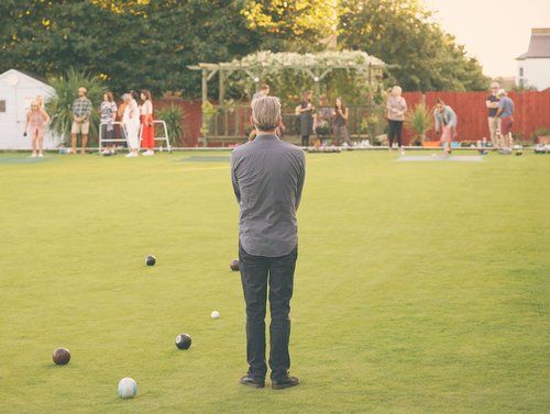 A person stands on a lush green lawn bowling green watching others play a game of bowls on a sunny day.