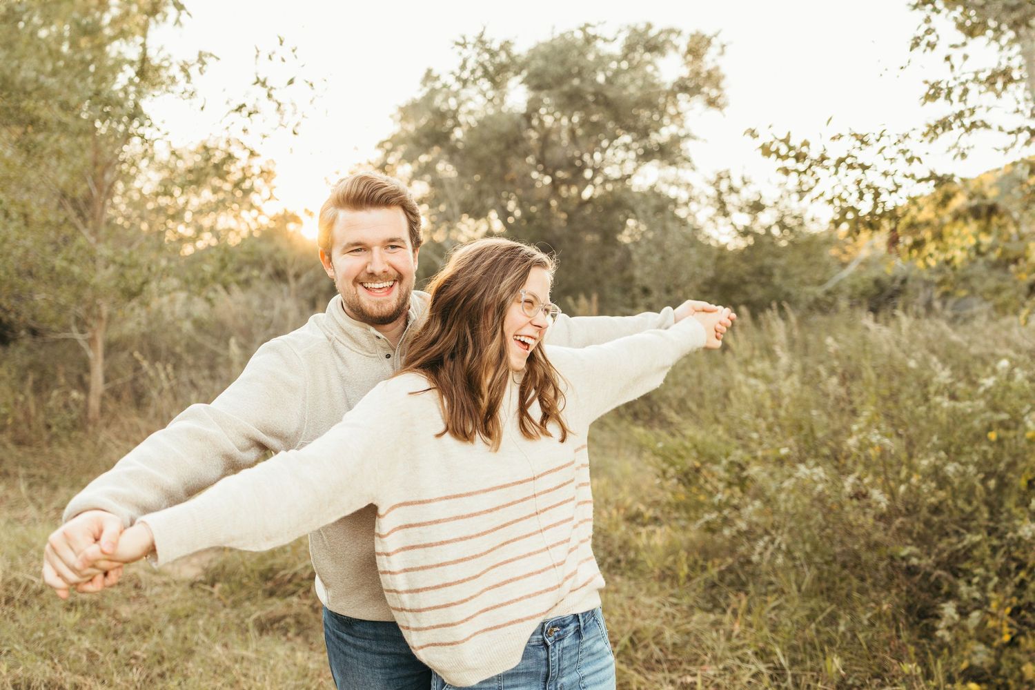 Happy couple embracing outdoors at golden hour, laughing joyfully in a sunlit field with autumn foliage in League City TX.