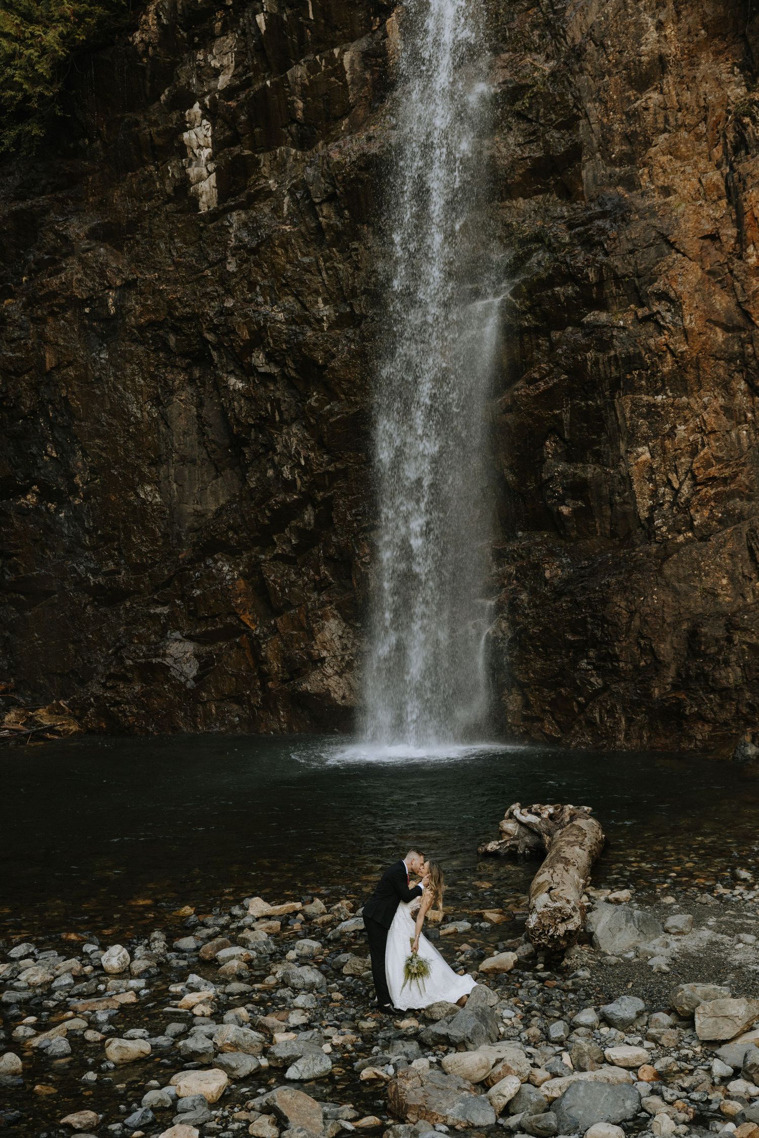 Bride and groom share a romantic kiss in front of a stunning waterfall surrounded by rocky terrain in Snoqualmie
