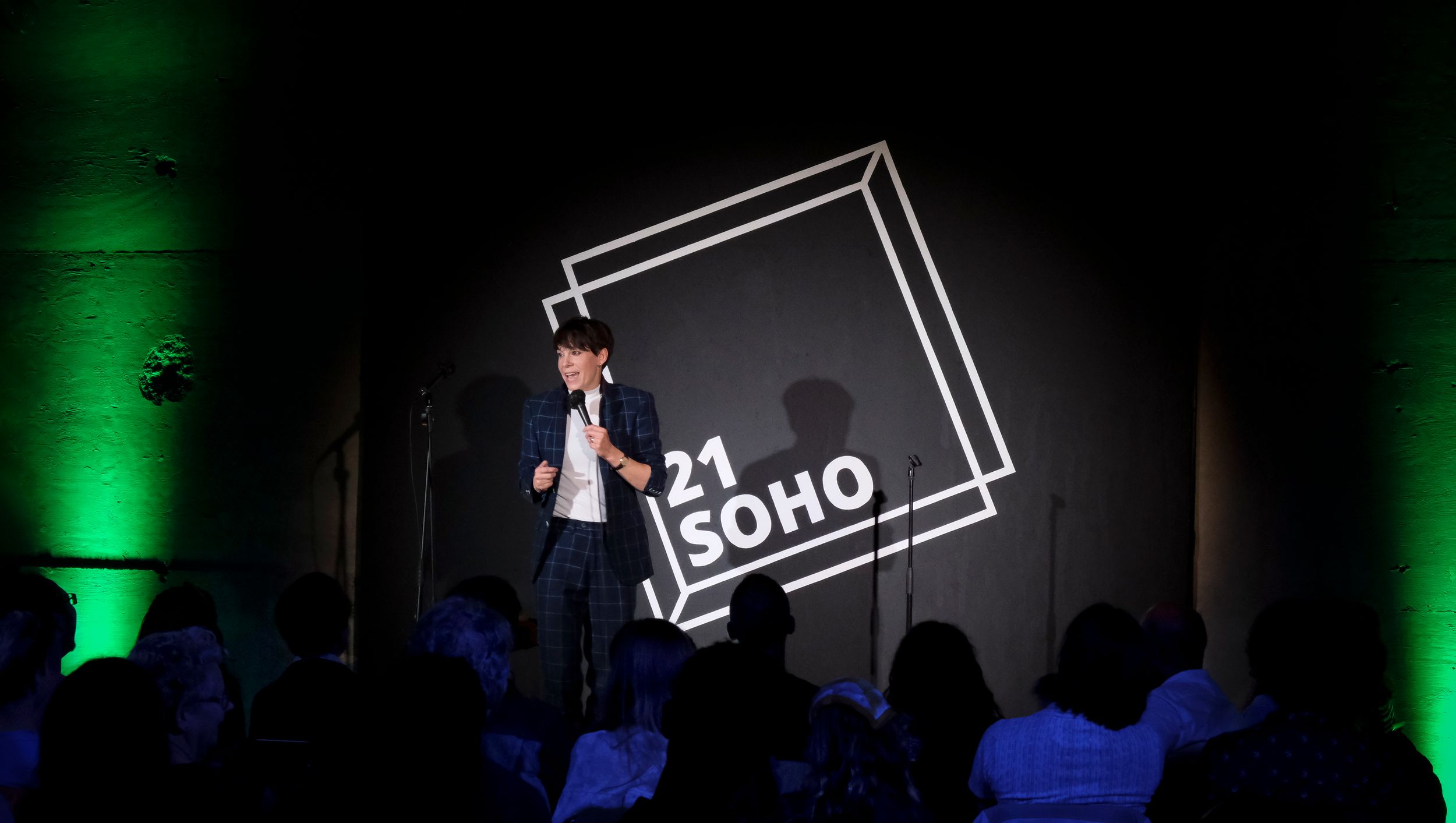 Speaker presenting on stage at 21 SOHO event venue with green lighting and audience silhouettes.