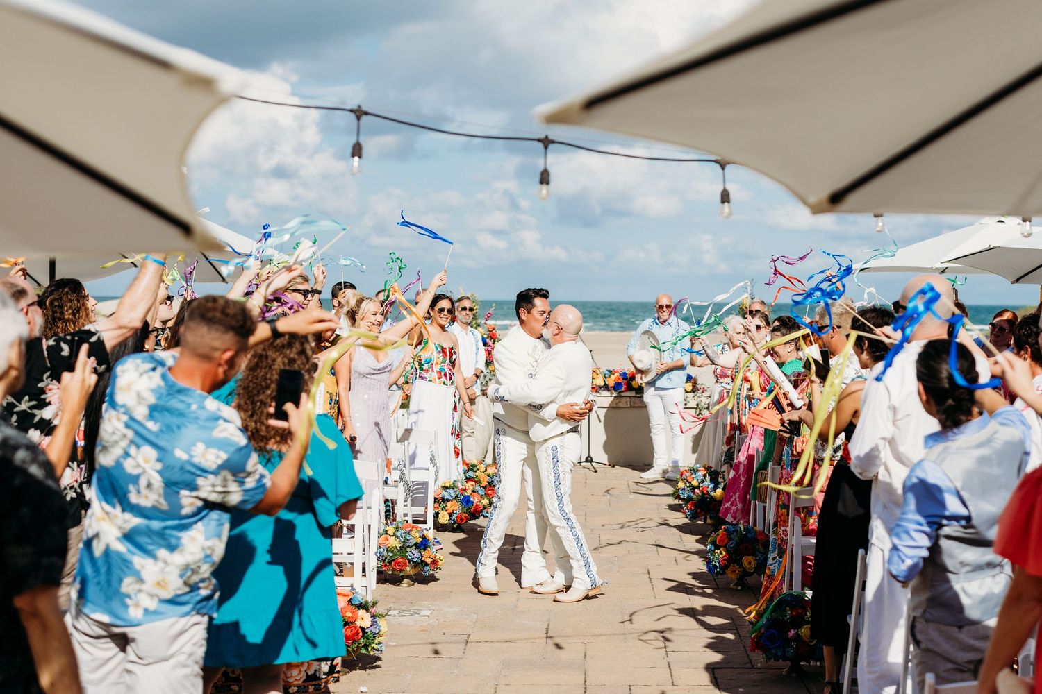 Joyful wedding couple walks through cheering guests waving colorful ribbons at outdoor beach reception.