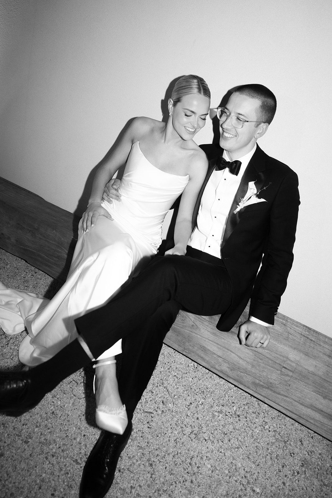 Black and white photo of a smiling bride and groom in formal attire sitting together on a bench at their wedding.