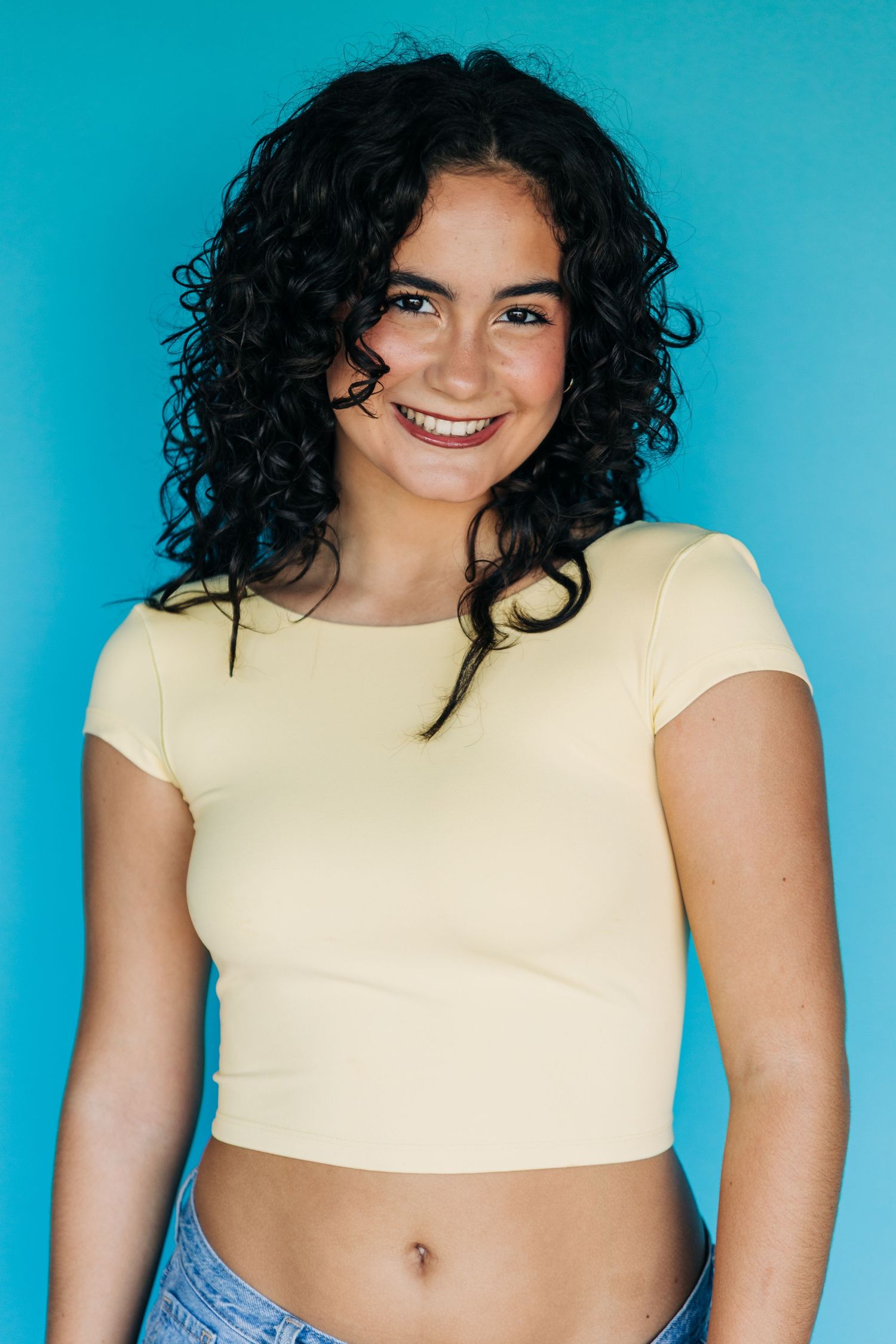 Smiling young woman with curly black hair wearing a yellow crop top against a bright blue background.