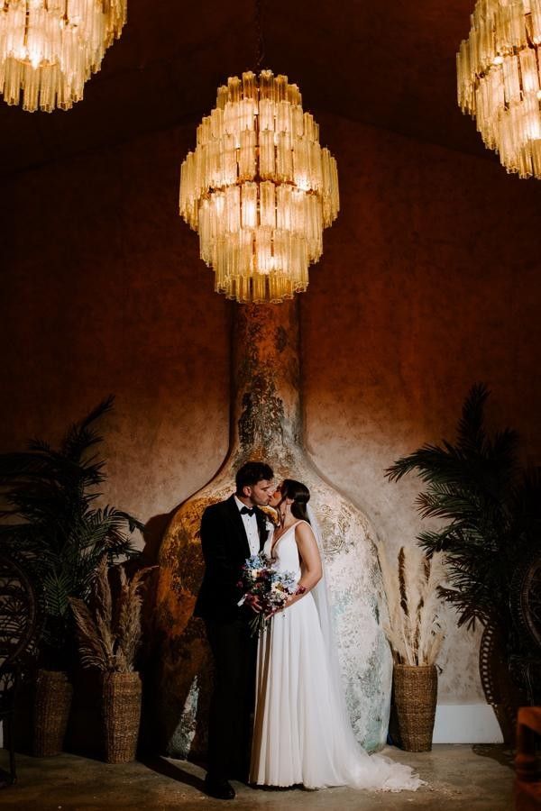 Newlyweds sharing a kiss beneath a chandelier as guests throw confetti
