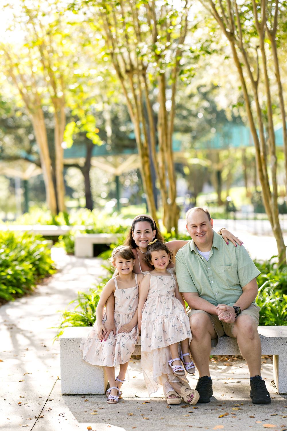 Happy family of four posing on a park bench surrounded by lush green trees on a sunny day.