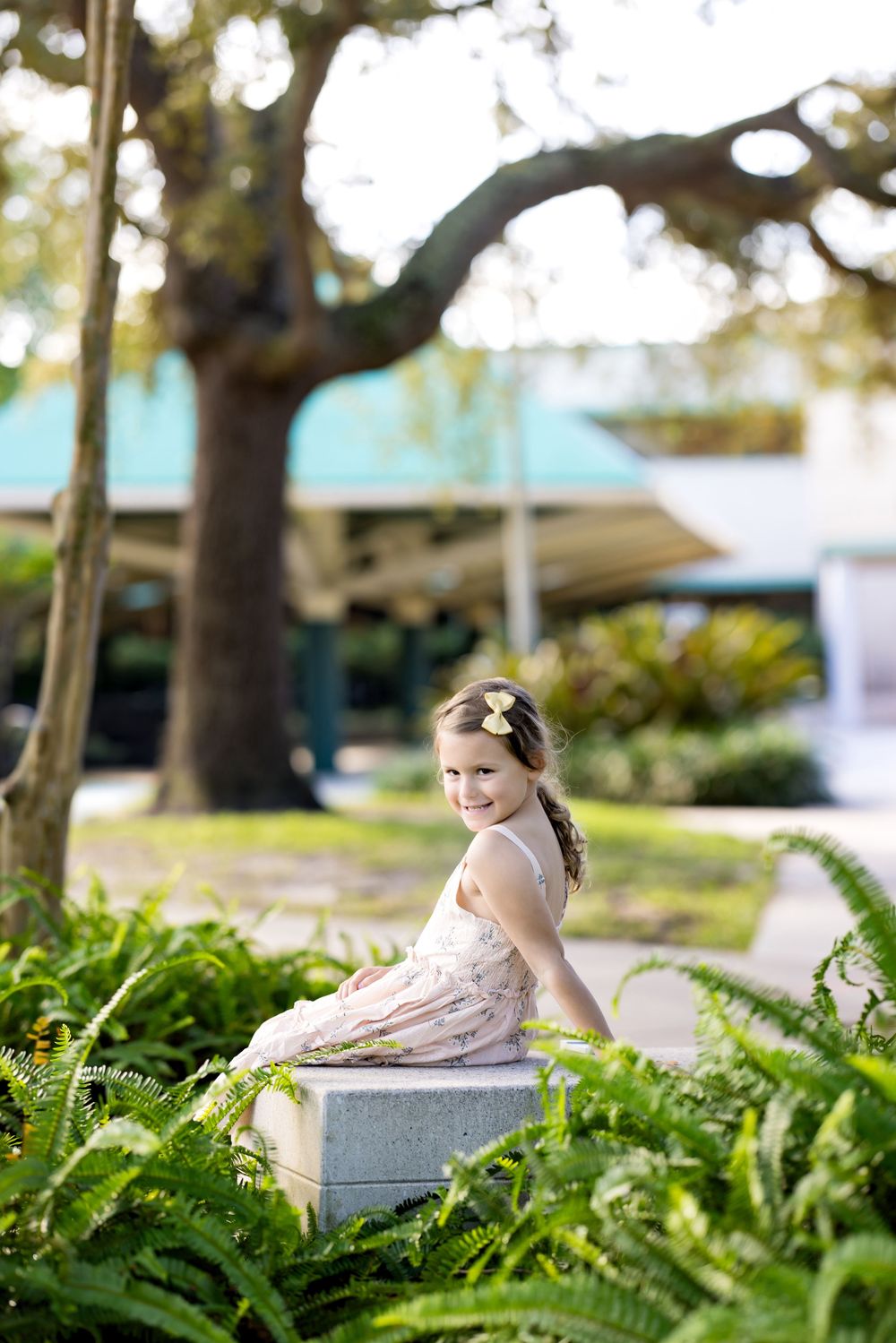 A smiling young girl in a floral dress sits on a stone planter surrounded by ferns in a sunny park setting.