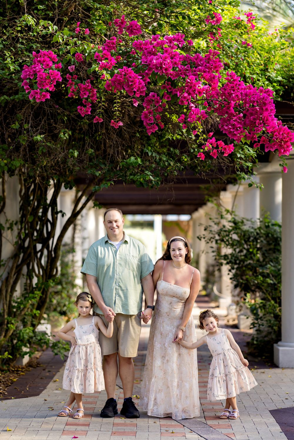 Family of four poses under vibrant pink bougainvillea on a brick pathway, parents holding hands with two young girls in matching dresses.