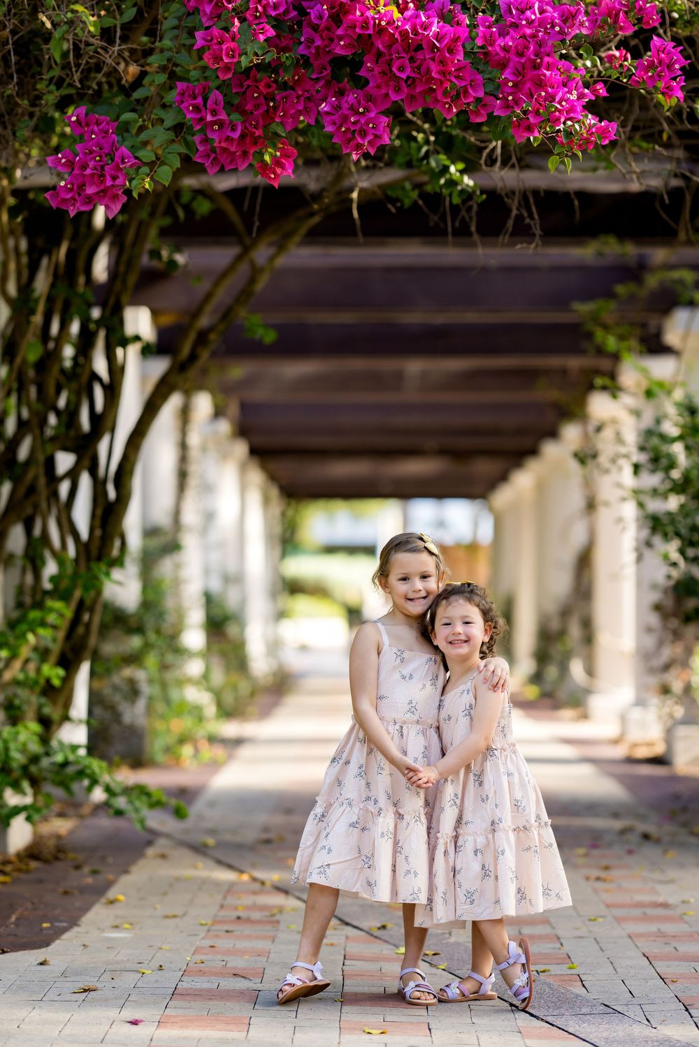 Two young girls in matching floral dresses pose under a bougainvillea-covered pergola on a brick pathway.