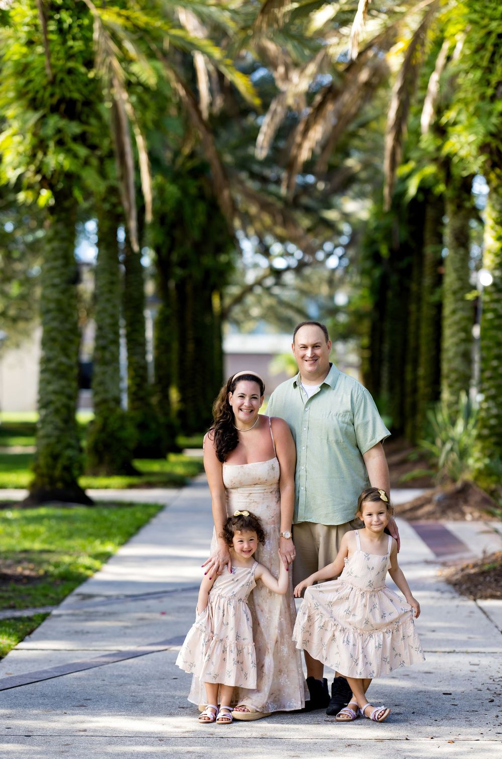 Family of four posing on a palm tree-lined walkway, parents with two young girls in matching dresses outdoors.
