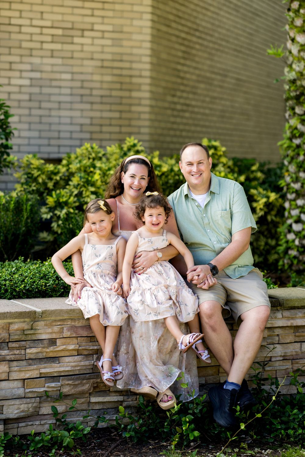 Happy family of four poses on a stone wall surrounded by lush greenery, parents with two young daughters in matching dresses.