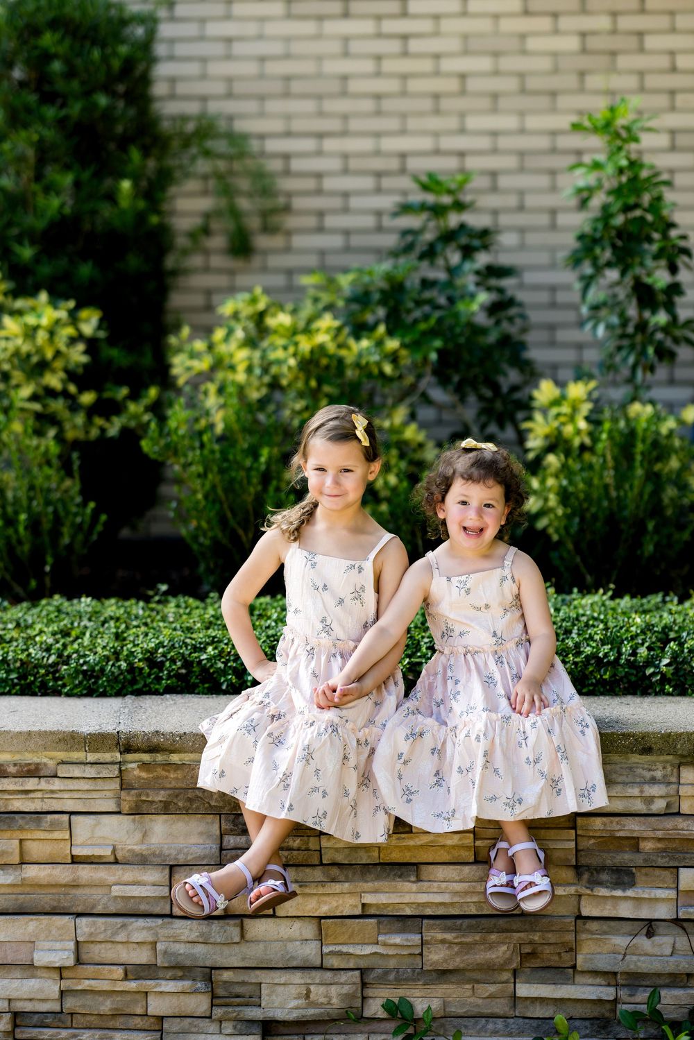 Two young girls in matching floral dresses sit together on a stone wall surrounded by lush green garden shrubs.