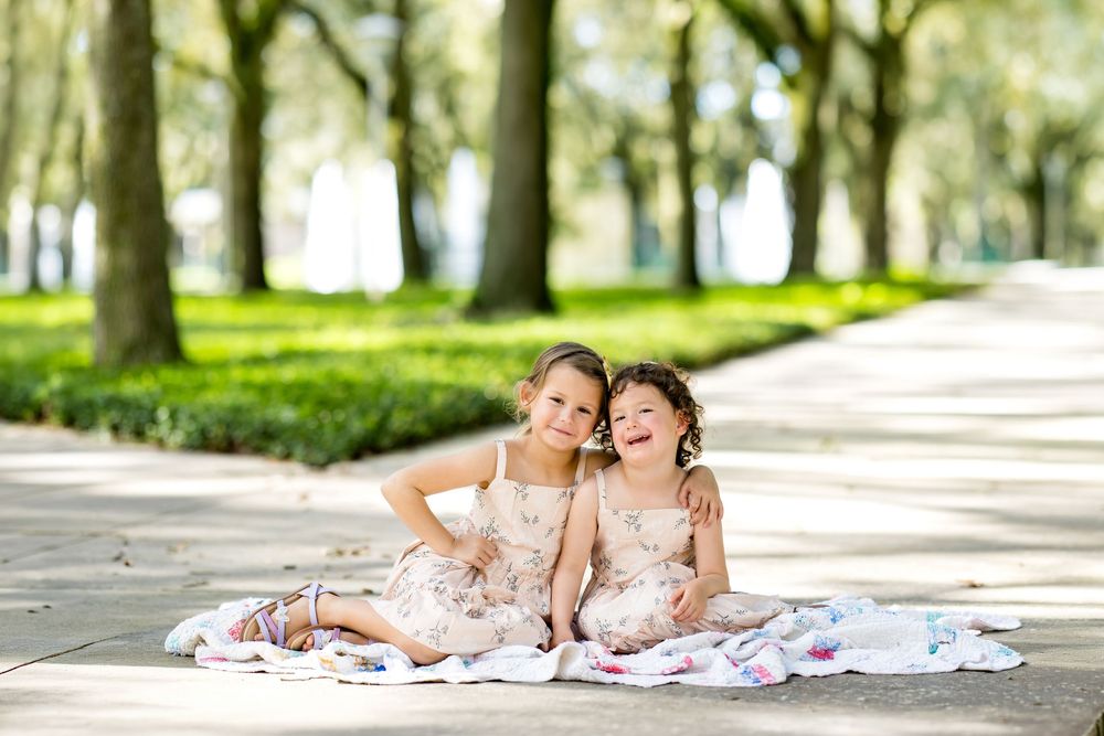 Two young girls in matching floral dresses sit together on a blanket, laughing outdoors among tall trees.