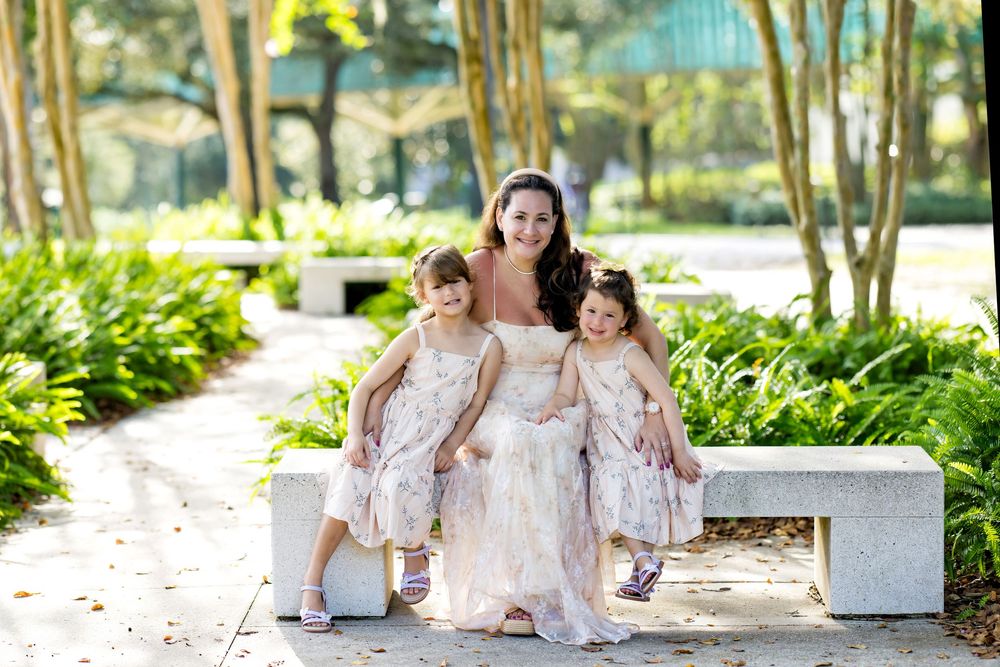 Bride in white gown poses on stone bench with two young flower girls in floral dresses outdoors.