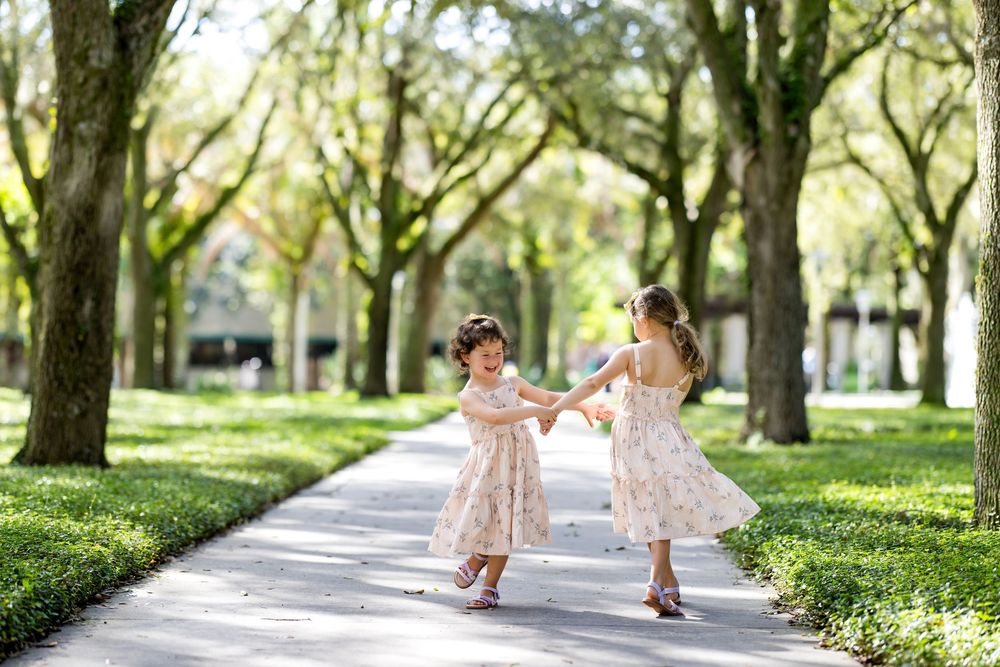 Two young girls in matching floral dresses hold hands and dance on a tree-lined pathway surrounded by lush greenery.