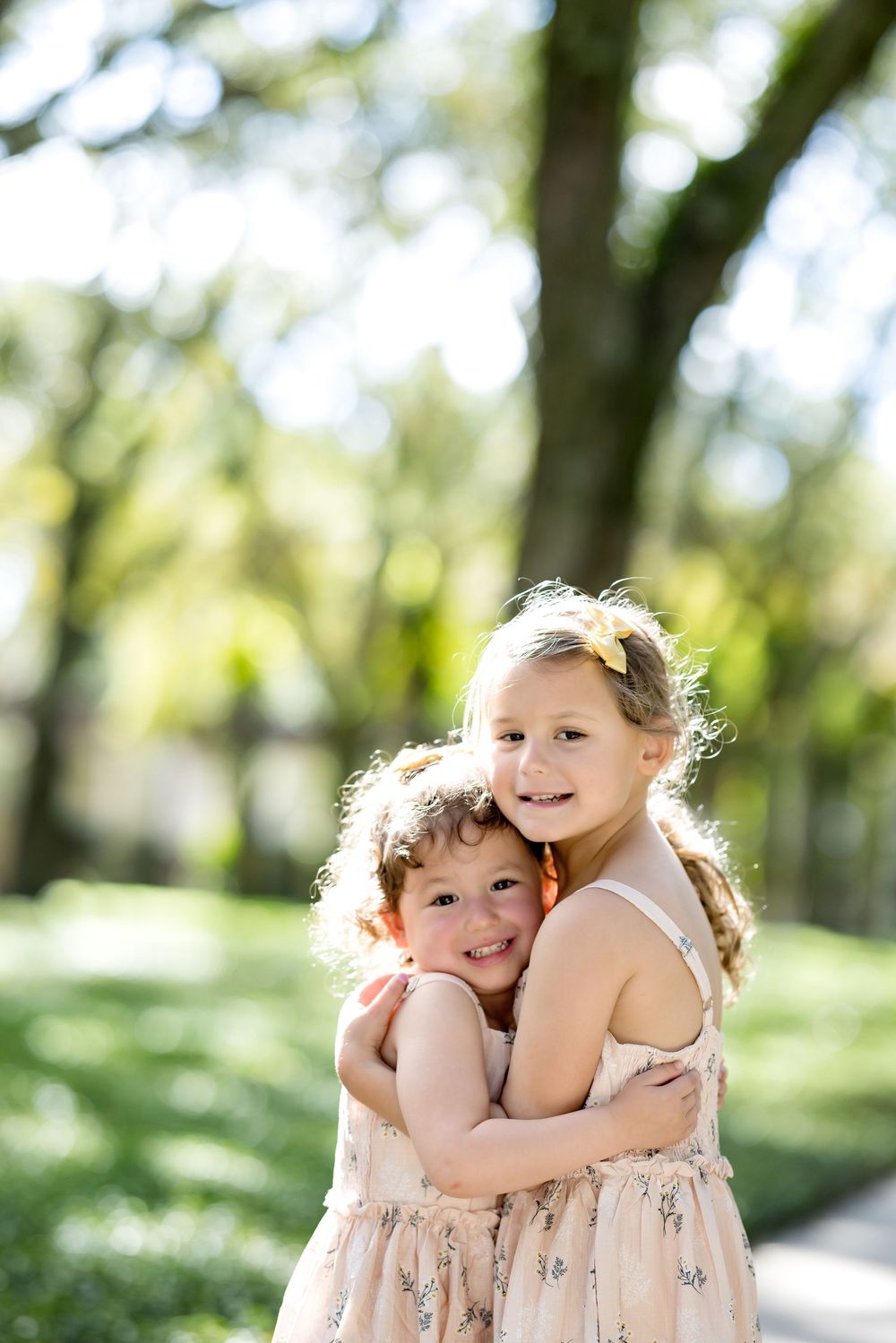 Two young sisters in matching floral dresses share a warm hug outdoors in a sunny green park.