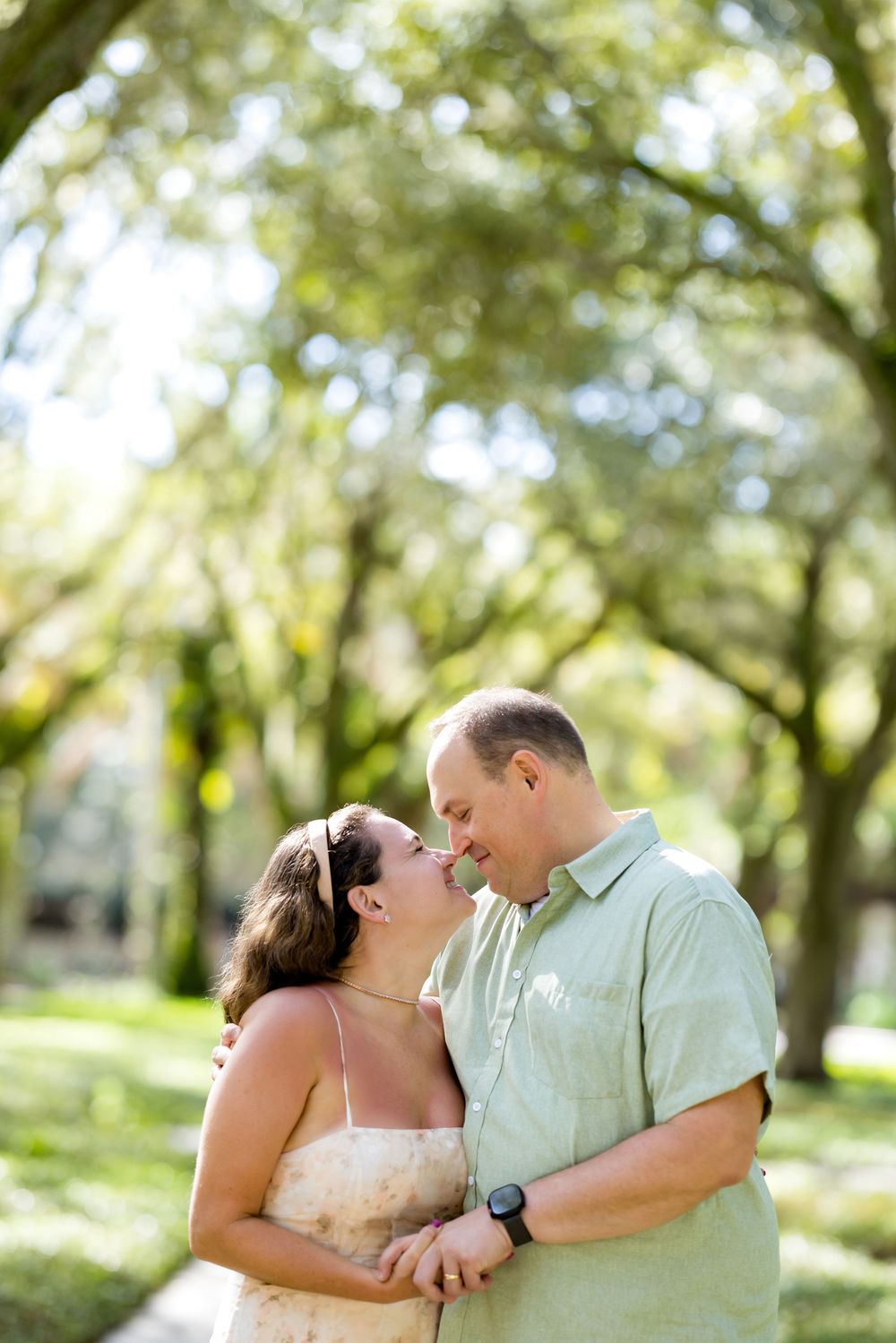 Couple sharing a romantic almost-kiss outdoors under a canopy of green trees in a sun-dappled park setting.
