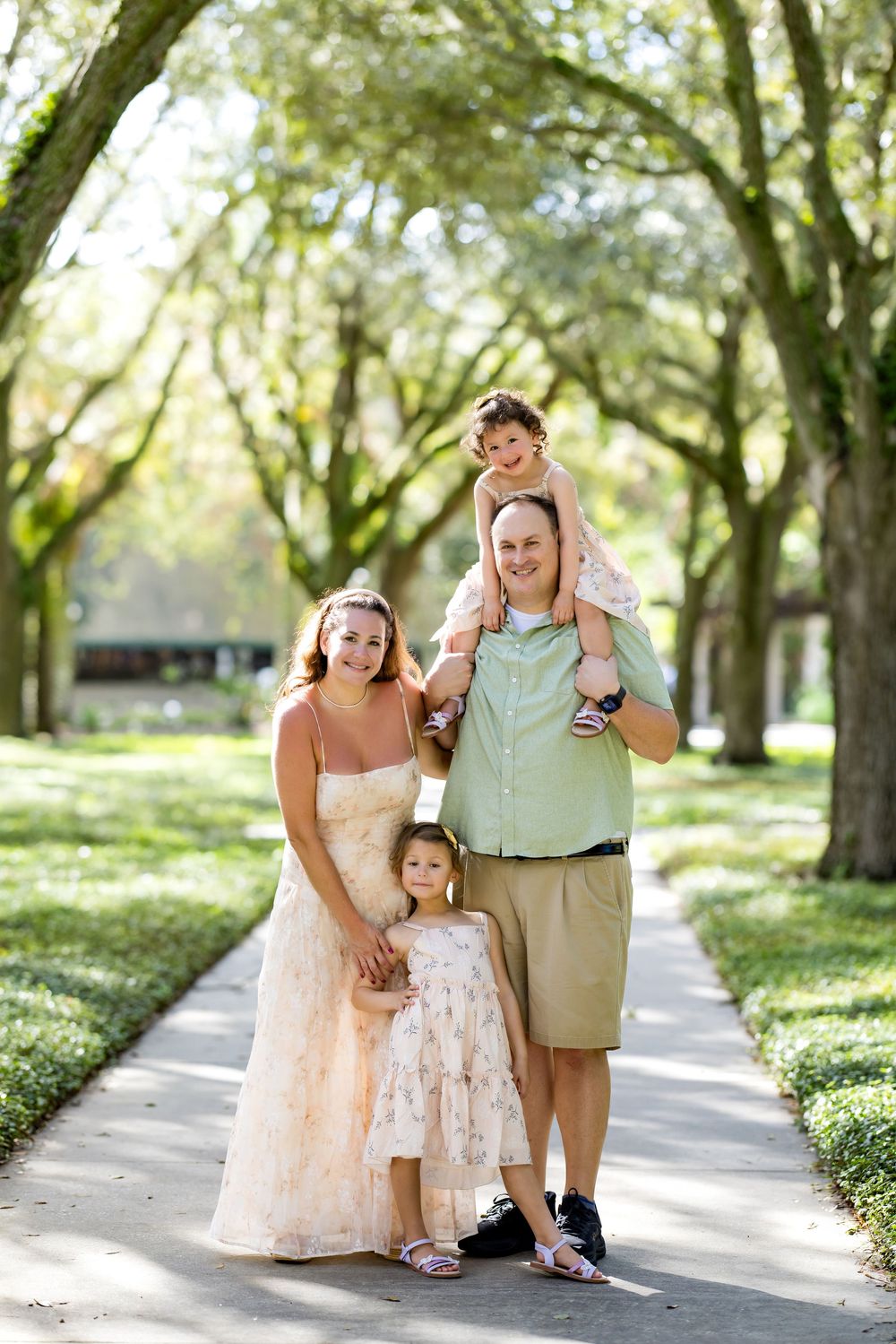 Family of four poses on a tree-lined path, parents with two young children, in a sunny park setting.