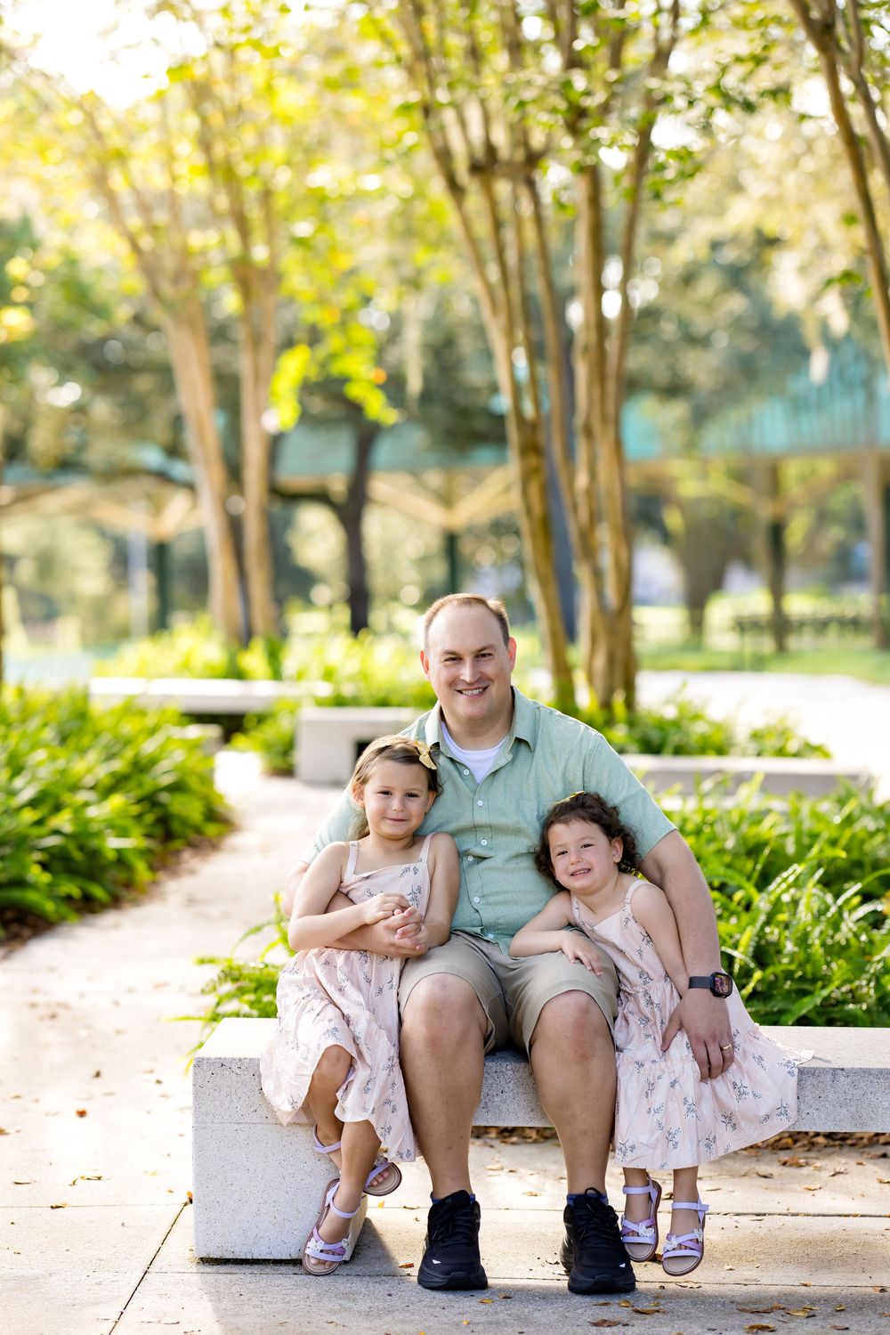 A smiling father sits on a stone bench in a sunny park with his two young daughters in floral dresses.