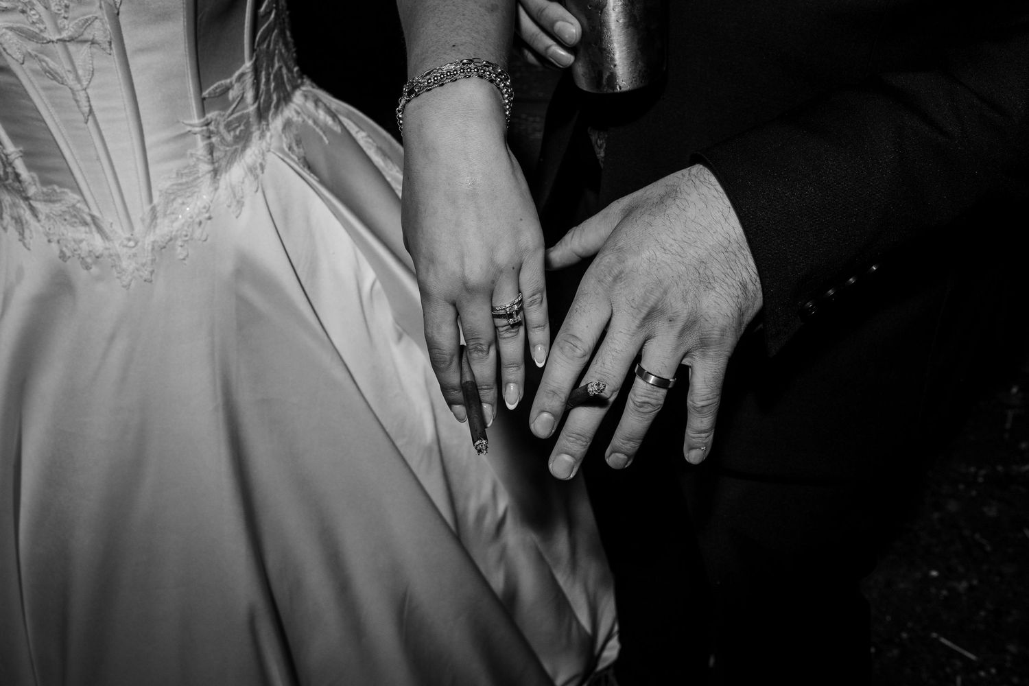 Black and white close-up of a bride and groom's hands showing their wedding rings, with the bride's lace bodice and gown beside them.