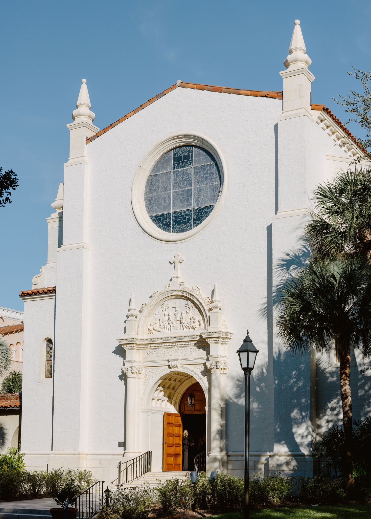 White Spanish Colonial church facade with rose window, ornate doorway, cross, and palm trees under blue sky.