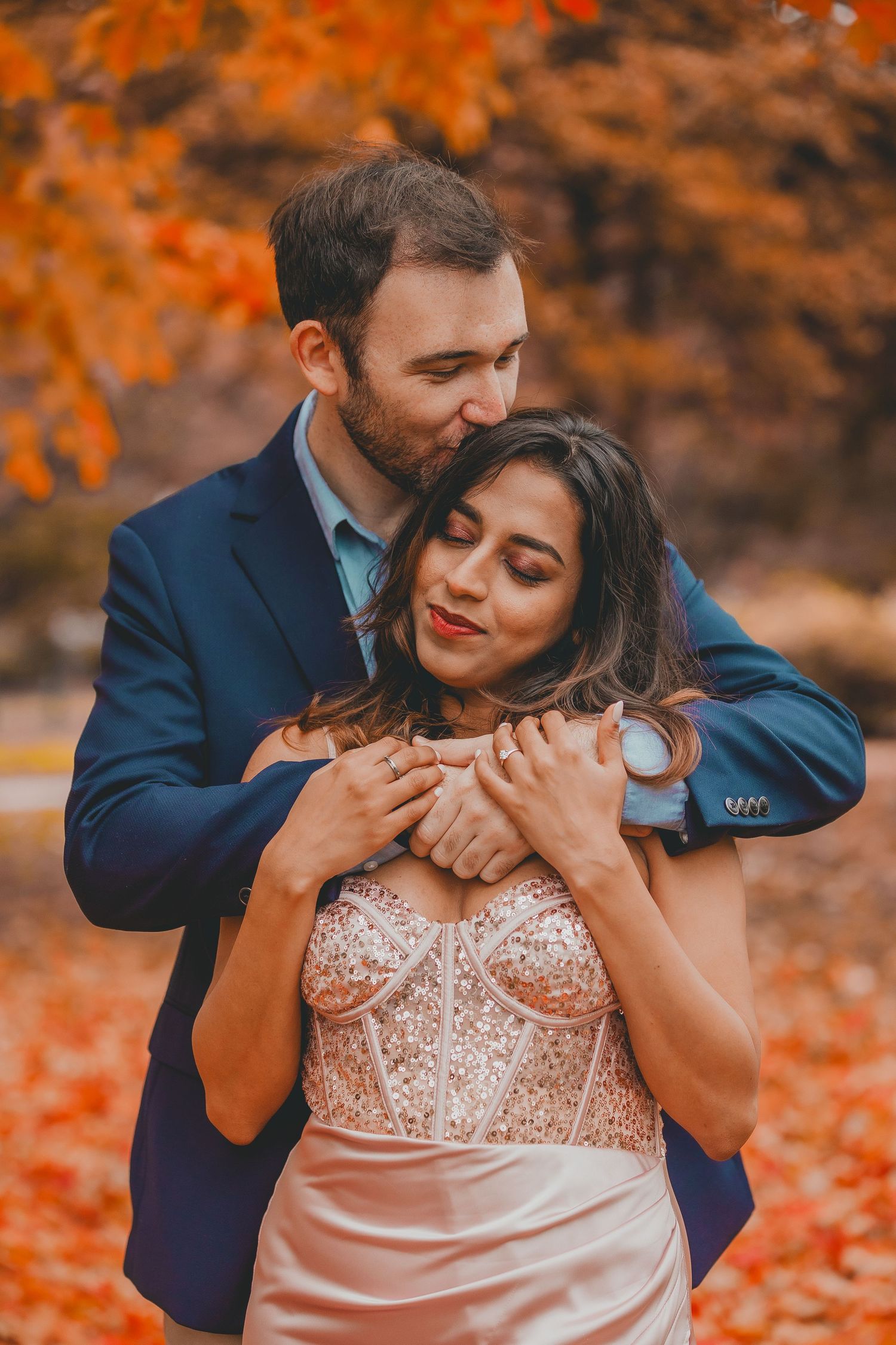 Bearded man in a navy blazer kisses a smiling woman's forehead as he hugs her from behind among vibrant orange autumn leaves.