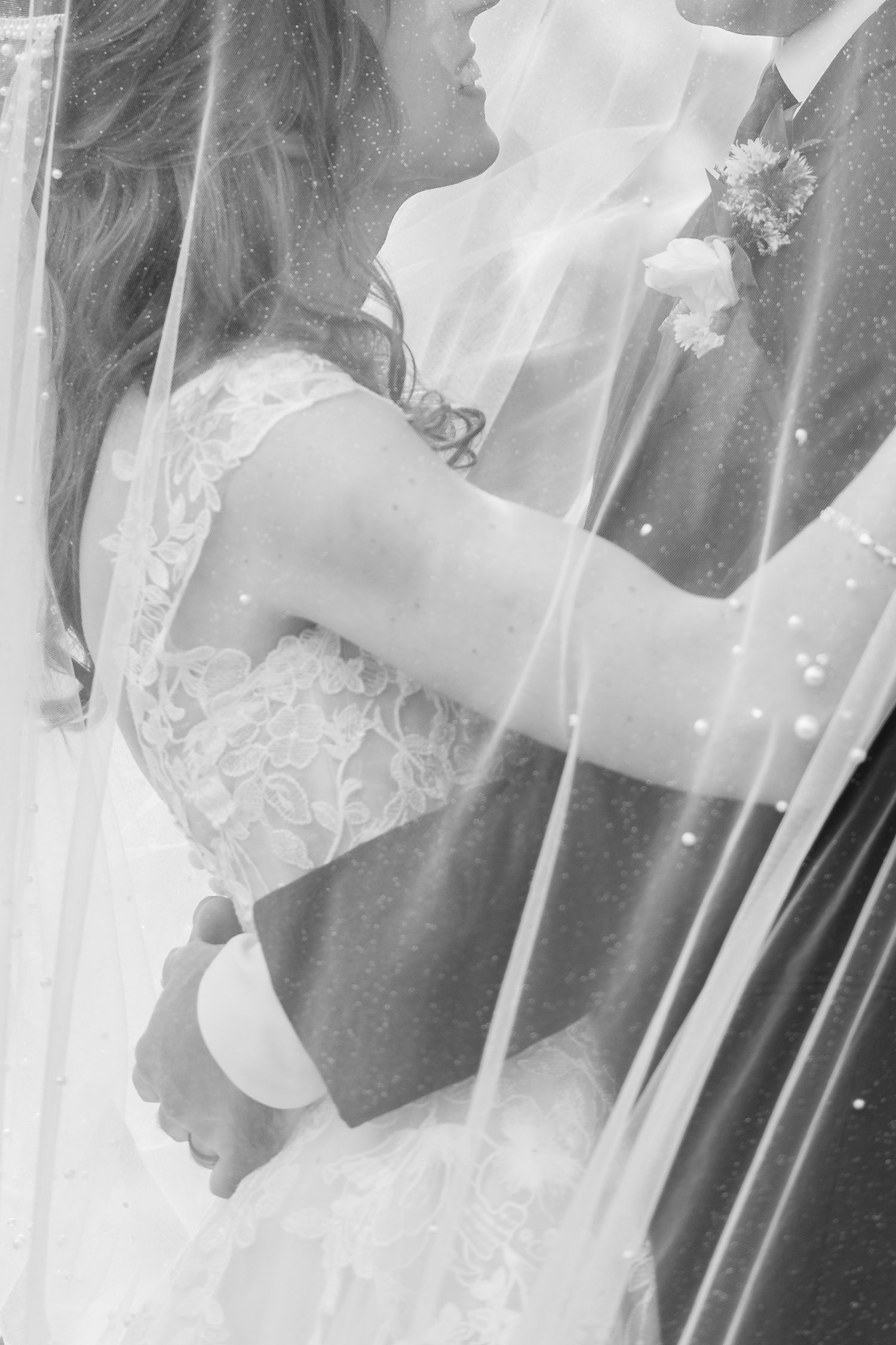 Black and white photo of a bride in a lace gown embracing a groom with a boutonniere, both seen through a sheer veil.