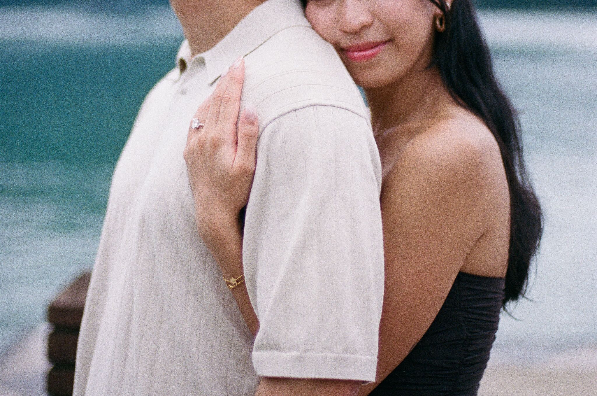 Couple embracing near turquoise ocean water, woman in black off-shoulder top smiling against man's shoulder.