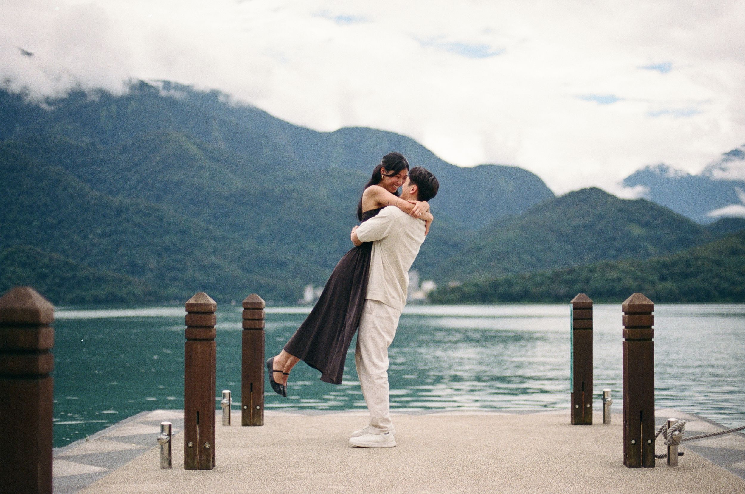 Couple embracing on a lakeside dock with misty mountains and turquoise water in the background.